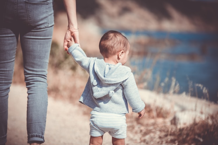 A woman holding the hand of her child outdoors, in a dry environment on a dusty trail, with a pond in the background. The woman and child have their backs facing us.
