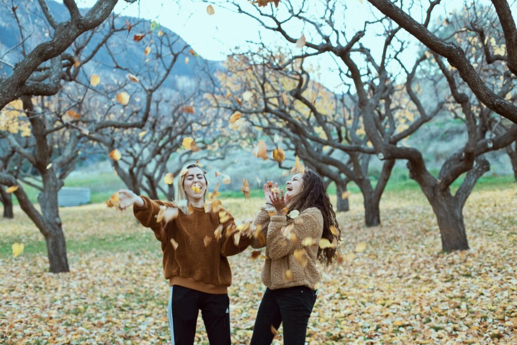 Two girl friends in park throwing yellow and orange leaves in the air in fall. Featuring leafless trees and hills in the background.